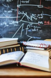 Stationery and notebooks scattered on a wooden desk ready for study near a chalkboard filled with notes. Generative AI